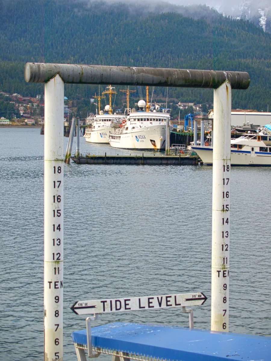 A tide-level staff gauge on a dock in Juneau, Southeast Alaska, with NOAA vessels moored in the background